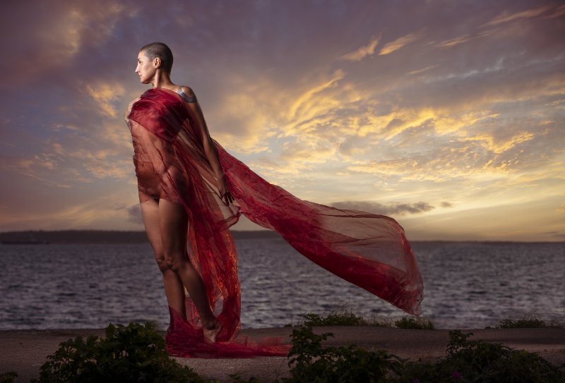 Woman posing in red sheer fabric by the sea in sunset